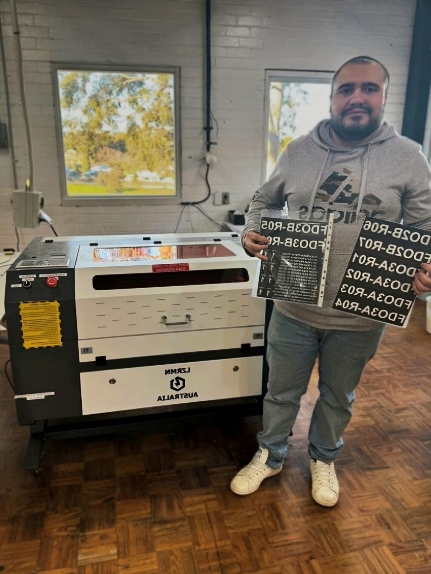 Andres is standing beside a laser cutting and engraving machine, showing off some recently made product.