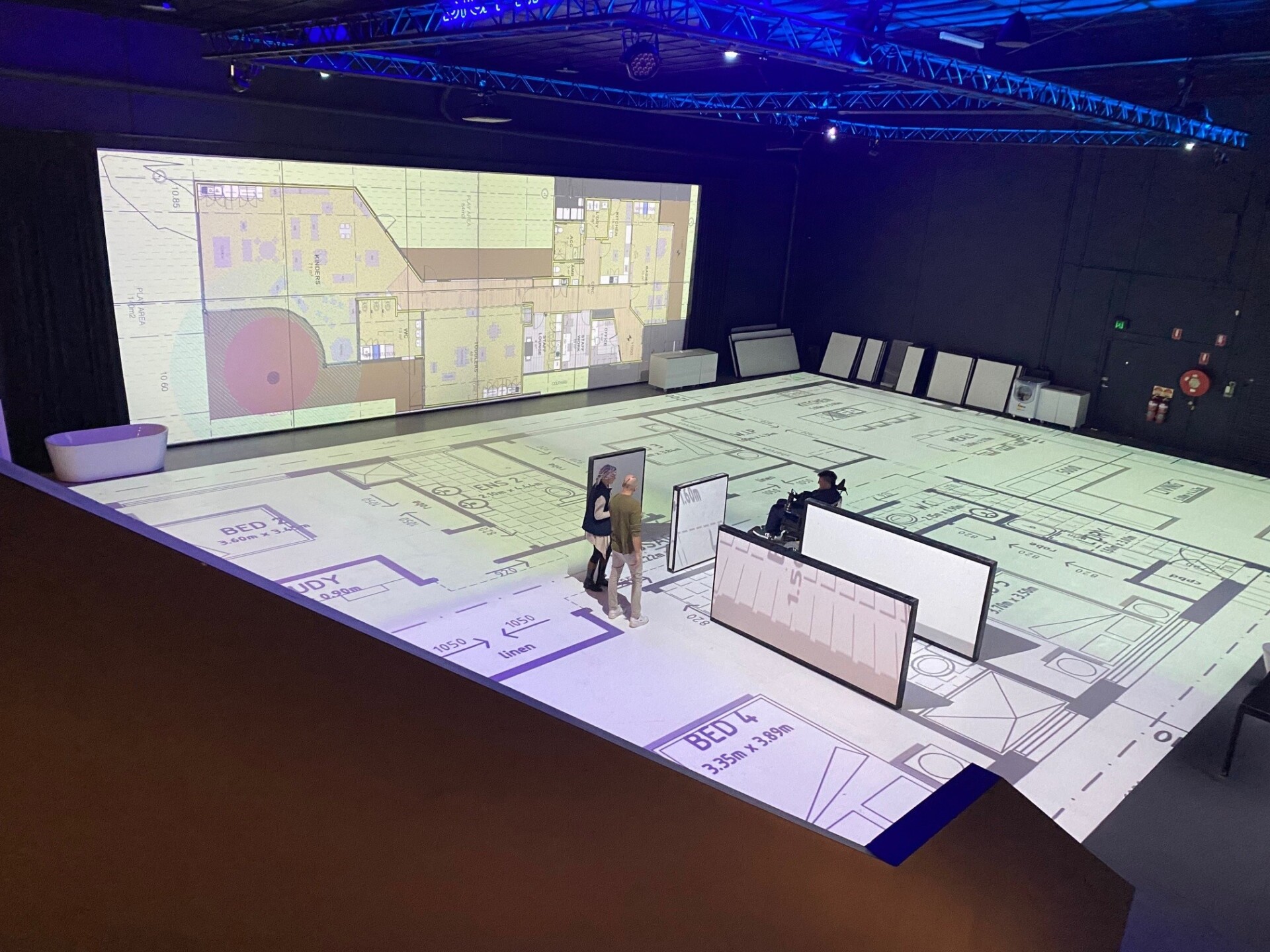 Mark uses a wheelchair. He goes through a full-size floor plan. This helps him understand space better before it is built. Two staff members watch him.