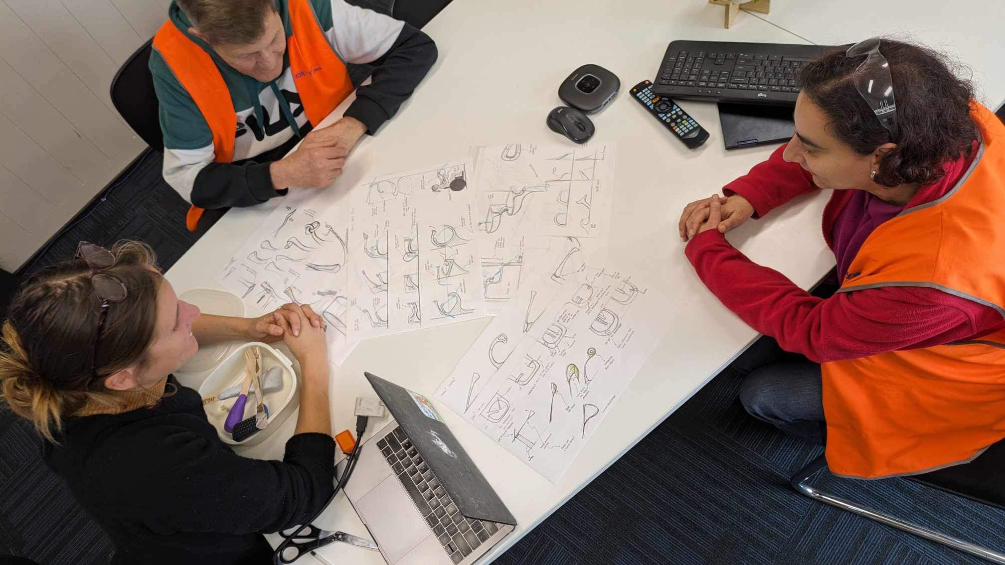 Swinburne University student is sitting at a table covered in tap design sketches with 2 Ability Works’ employees.