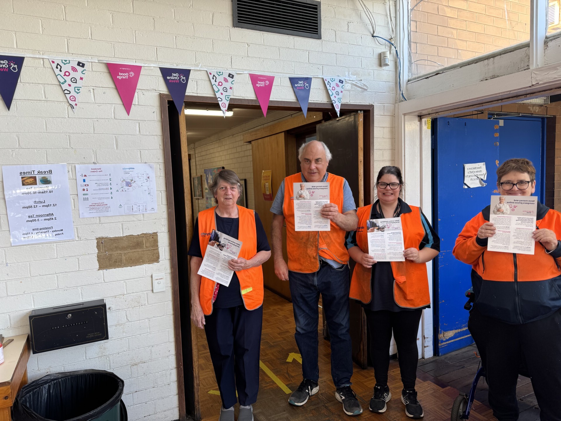 Four happy Ability Works employees standing under the bunting, holding up ‘Get Online Week’ flyers.