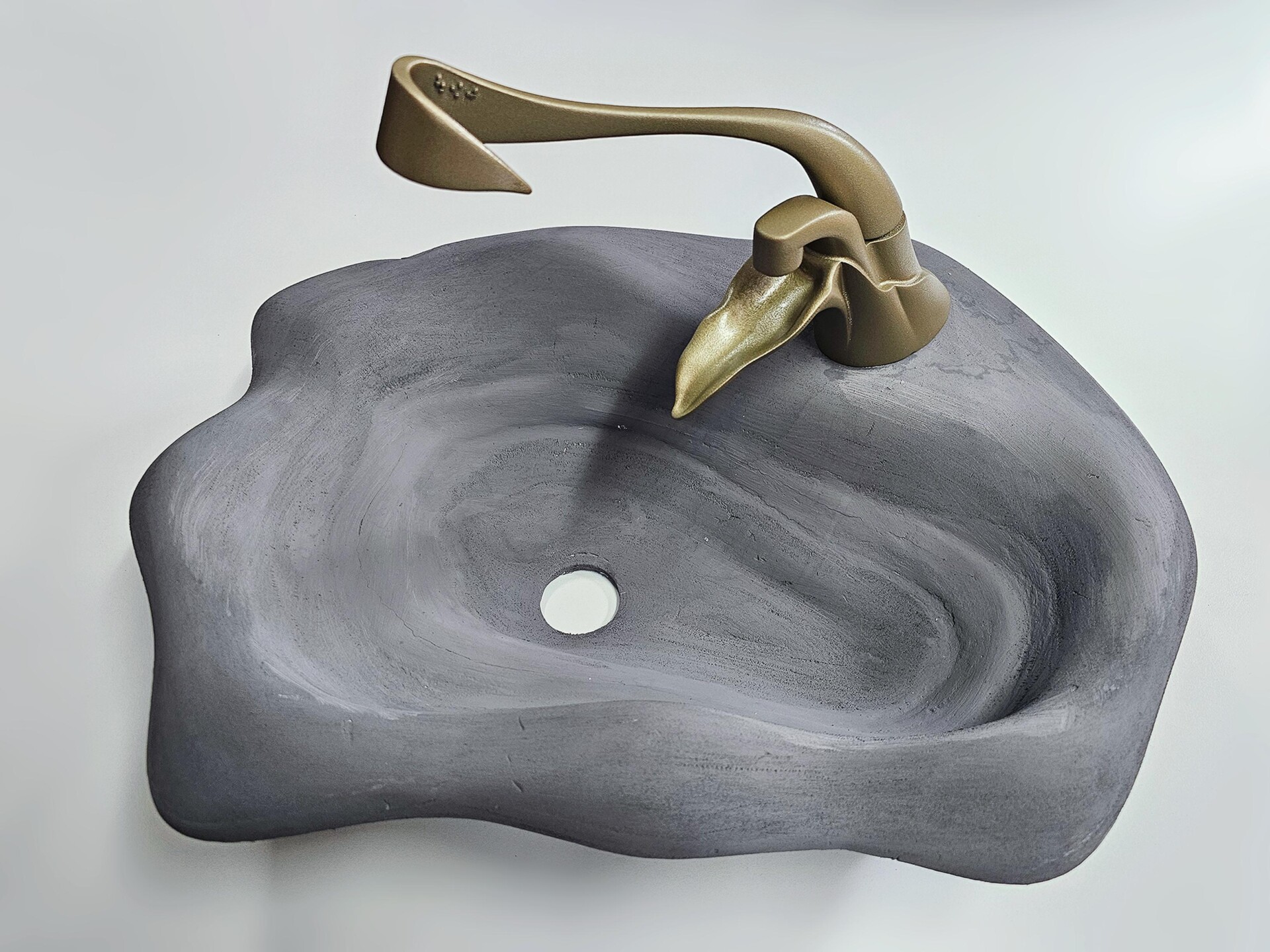 The tap is printed in a gold or brass colour and it has a rather natural shape with lots of curves, mimicking leaves. The handle has braille to indicate temperature and has a simple lever mechanism to start/stop the water flow. There is a leaf shaped structure below the faucet to assist the water to flow closer to the user. The basin is a natural grey colour, it appears to be made of stone and has a smooth, irregular shape as if it were real stone naturally eroded by water over time